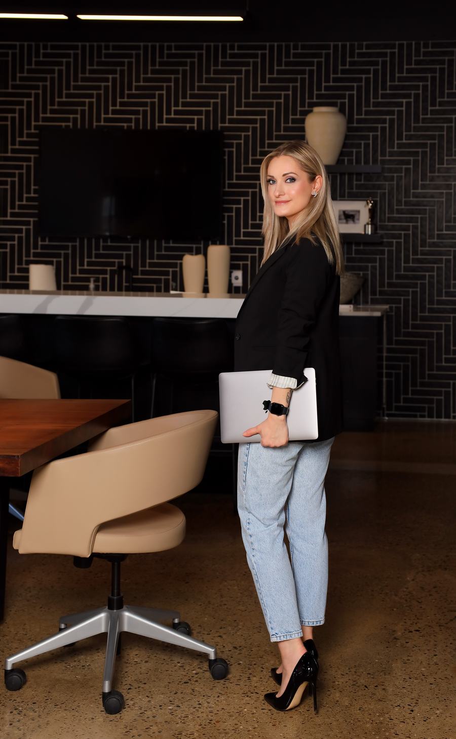 A blazer and denim in a dark herringbone office, laptop under one arm