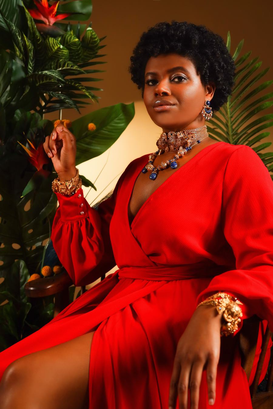 A red wrap dress and gold cuffs, seated among palms
