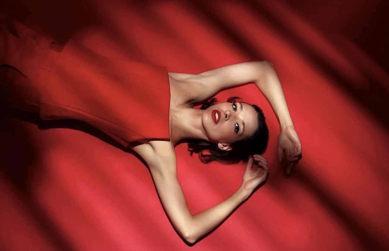 Overhead — a figure on red drapery, arms framing the face