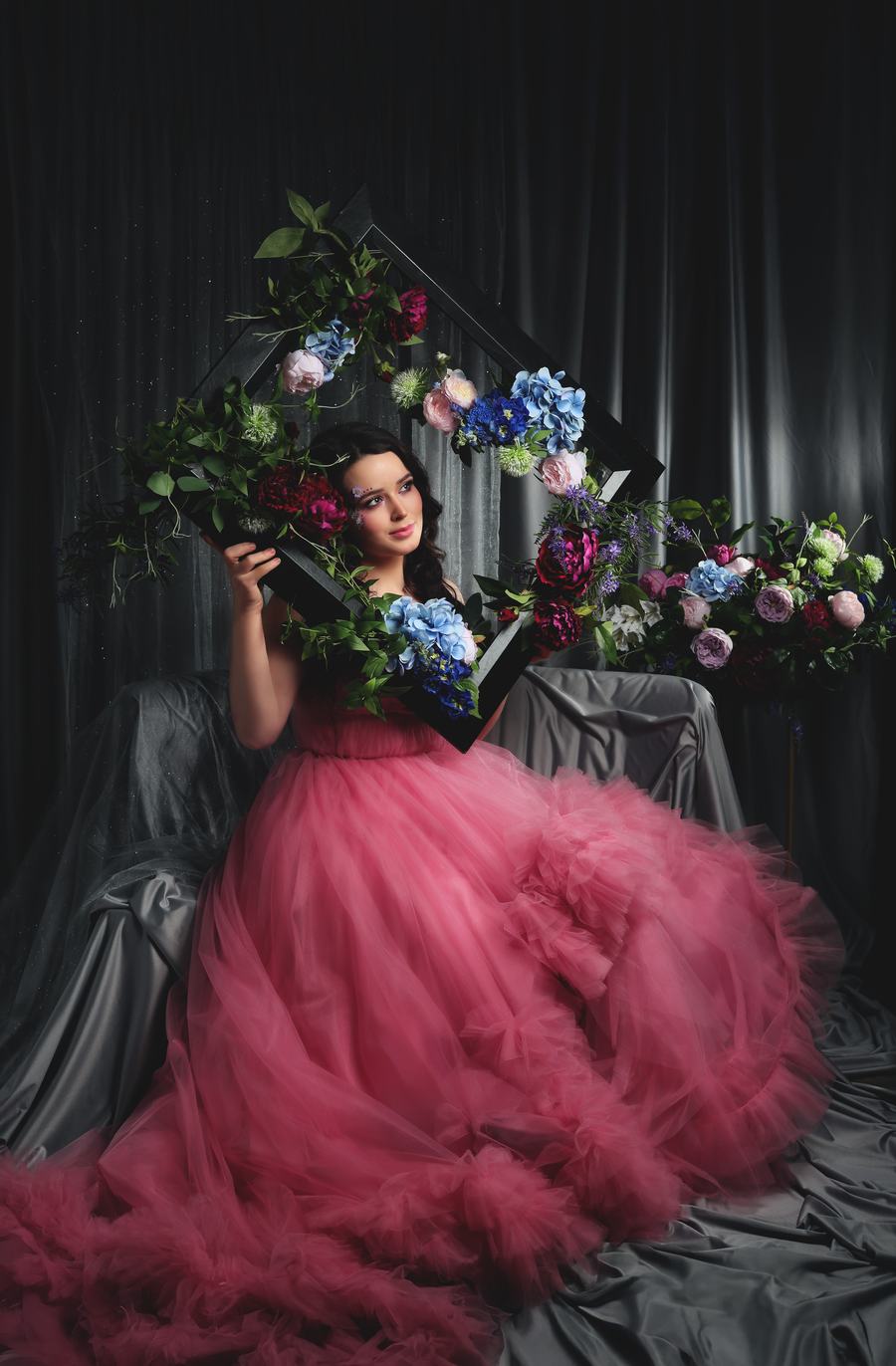 A rose-pink tulle gown, framed in flowers before dark drapery